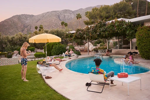 Outdoor pool party at sunset with group of people relaxing and swimming near a mid-century modern house, and mountain backdrop.