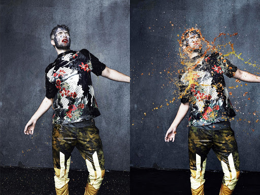Male model in a floral shirt with camouflage pants splattered with liquid, photographed in a studio against a gray textured background.