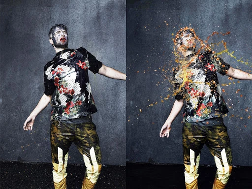 Male model in a floral shirt with camouflage pants splattered with liquid, photographed in a studio against a gray textured background.