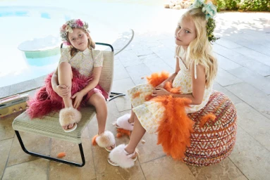 Two young girls wearing floral crowns and fluffy slides seated outdoors near a swimming pool with set design by Ariana Nakata.