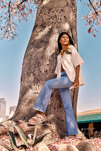 A woman in denim flares and a cream-colored top poses by a massive tree with budding flowers against a city skyline backdrop.