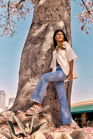 A woman in denim flares and a cream-colored top poses by a massive tree with budding flowers against a city skyline backdrop.