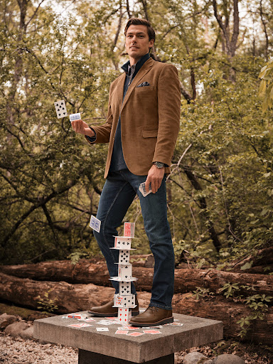 Full shot of a man balancing a card tower outdoors in a brown blazer, dark jeans, and brown leather dress shoes, surrounded by trees.