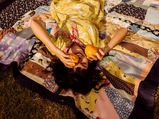A woman in a yellow dress lies on a colorful patchwork quilt outdoors, holding two oranges in her hands with a joyful expression.