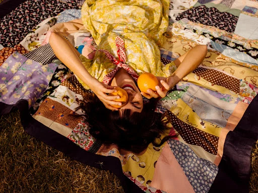 A woman in a yellow dress lies on a colorful patchwork quilt outdoors, holding two oranges in her hands with a joyful expression.
