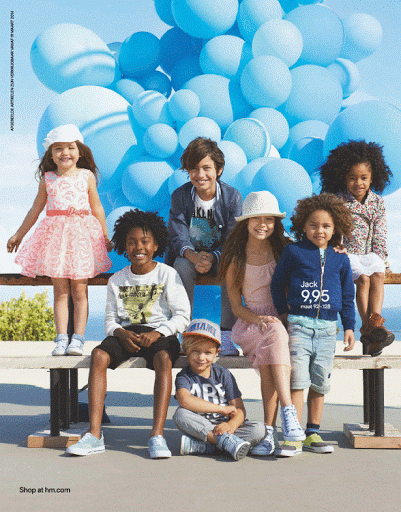 Group of seven children of diverse ethnicities are sitting and posing on a bench in front of many light blue balloons and a light blue sky.