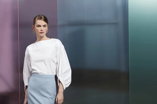 A poised woman with blonde hair, wearing a white blouse and blue pencil skirt, stands in front of an abstract colorful background.