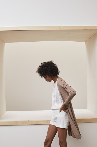 A slim African-American woman poses indoors with a hand on her hip, wearing a long cardigan, white top, and white shorts.