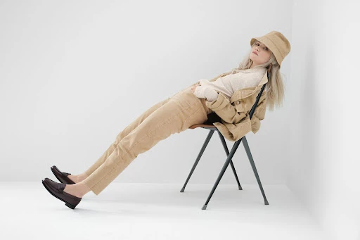 A woman with fair hair lounges on a chair in a beige bucket hat, jacket, top, and pants, complemented by brown shoes against a white background.