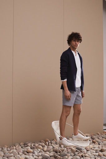 A male model in a navy blazer, white t-shirt, and patterned shorts poses while standing on a chair in a fashion shoot.