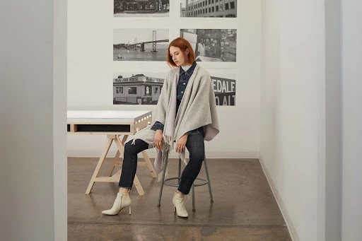 A red-haired woman wearing white heeled boots and a grey shawl sits on a stool indoors in front of black and white photographs.