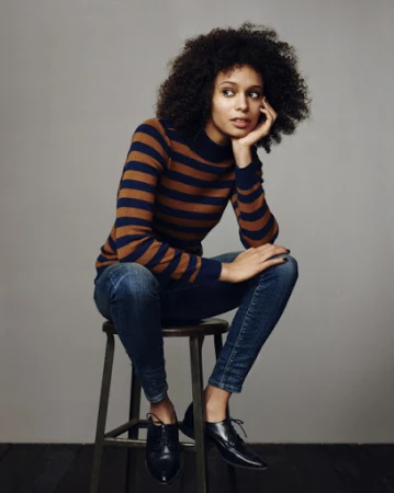Woman with curly dark hair wearing a striped sweater and jeans seated on a stool against a grey background