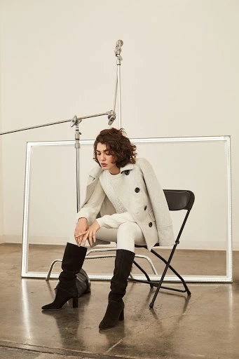 Woman in pea coat, sweater, and boots sits in front of a rectangular photo reflector in a studio, creating a stylish fashion portrait.