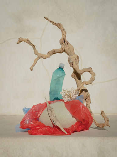 Still life featuring a plastic bottle, plastic bags, driftwood, and coral against a textured off-white wall. Studio lighting creates neutral tones.