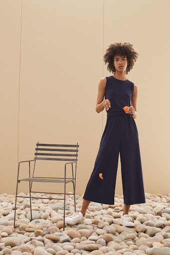 A woman wearing a sleeveless navy jumpsuit stands on a stone surface next to a metal chair against a light brown background.