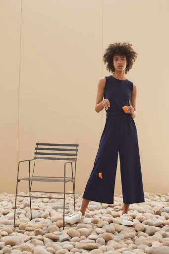 A woman wearing a sleeveless navy jumpsuit stands on a stone surface next to a metal chair against a light brown background.