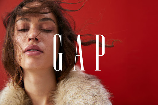 Brunette woman with closed eyes in a fur coat against a red backdrop with GAP text overlayed, fashion forward and modern.