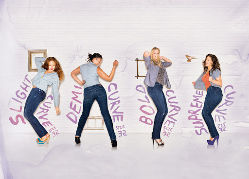 Four women in dark blue jeans dancing in front of a white wall with painted purple typography and small framed pictures