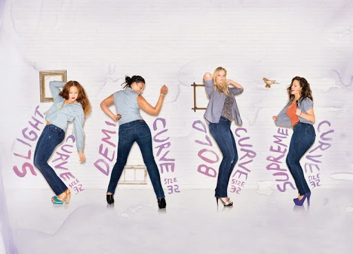 Four women in dark blue jeans dancing in front of a white wall with painted purple typography and small framed pictures