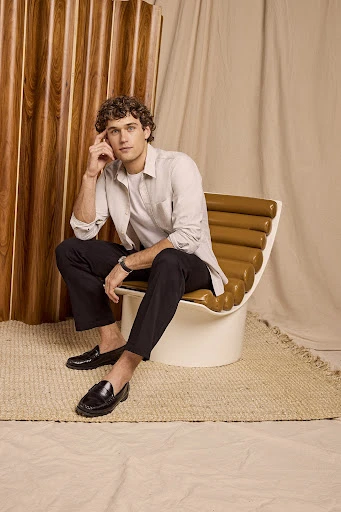 A young adult male with curly hair in a casual outfit sits on a brown chair in a studio setting with a relaxed pose, wearing loafers.