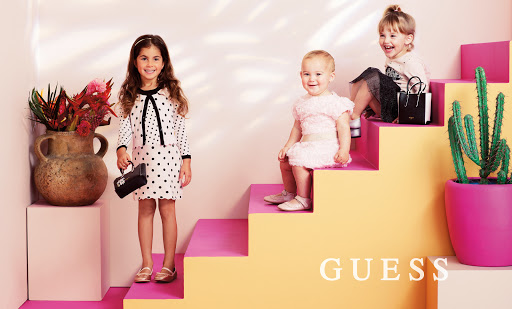 Three children pose on pink and yellow stairs in a studio. One girl holds a purse. Flowers and a cactus add a colorful touch.
