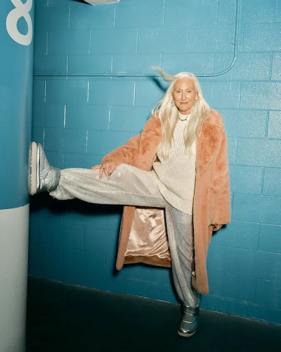 Elderly woman in peach coat and silver pants poses with leg raised against blue tiled wall. Stylish fashion portrait in urban setting.