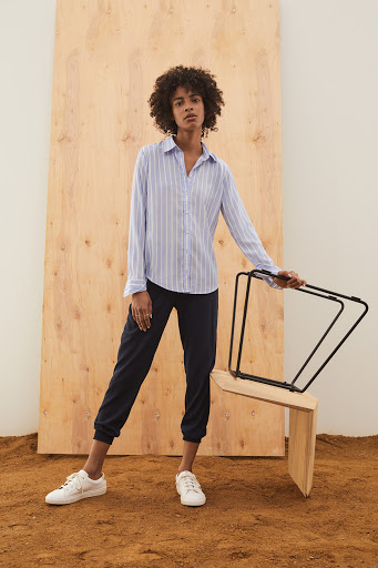 Adult female stands indoors wearing a striped button-down shirt, dark pants, and white sneakers, posed near a plywood wall and minimal chair.