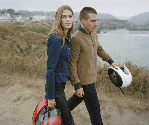 Man and woman walking outdoors carrying motorcycle helmets near water, styled by Chime Serra.