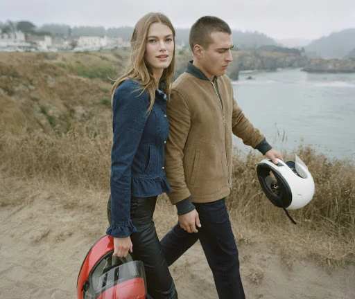 Man and woman walking outdoors carrying motorcycle helmets near water, styled by Chime Serra.