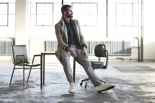 Bearded man in beige coat seated on a table in an industrial studio styled by Chime Serra
