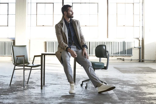 Bearded man in beige coat seated on a table in an industrial studio styled by Chime Serra