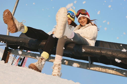 Two young girls in winter clothes riding a ski lift, smiling at the camera with a bright blue sky and snowy mountain visible.