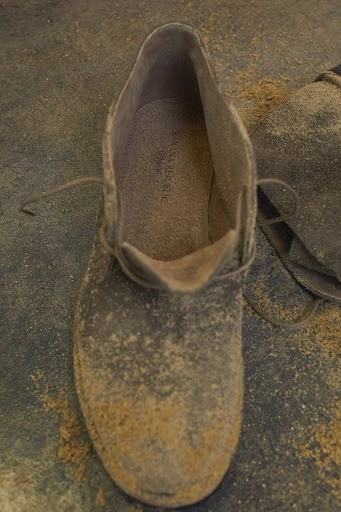 Close-up of a grey suede chukka boot with laces, covered in light-brown dust, sitting on a matching dusty surface.