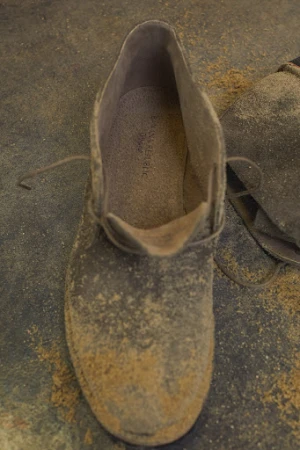 Close-up of a grey suede chukka boot with laces, covered in light-brown dust, sitting on a matching dusty surface.