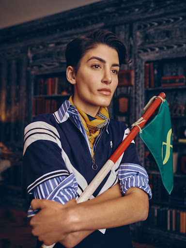 Portrait of an adult woman indoors, posing with a green flag with gold detail, wearing a striped collared shirt and patterned neck scarf.