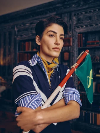 Portrait of an adult woman indoors, posing with a green flag with gold detail, wearing a striped collared shirt and patterned neck scarf.