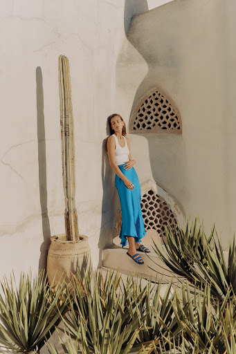 A woman poses in a white tank top and blue skirt on a textured white surface with a cactus and succulents in an outdoor setting.