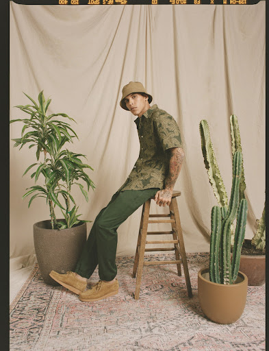 Man in patterned shirt and bucket hat poses seated on stool surrounded by potted plants in a neutral-toned photo studio setting.