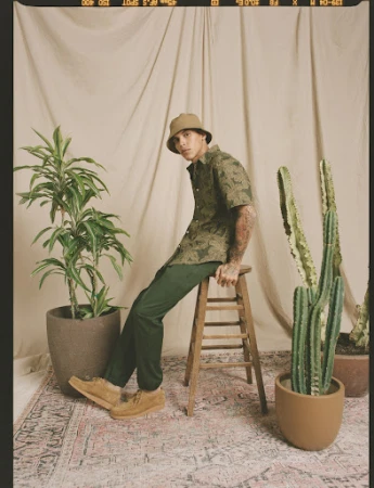 Man in patterned shirt and bucket hat poses seated on stool surrounded by potted plants in a neutral-toned photo studio setting.