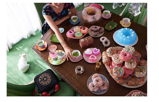 Overhead shot of woman at table with colorful donuts, cakes, and teacups, set indoors with pastel green carpet and a retro black telephone.