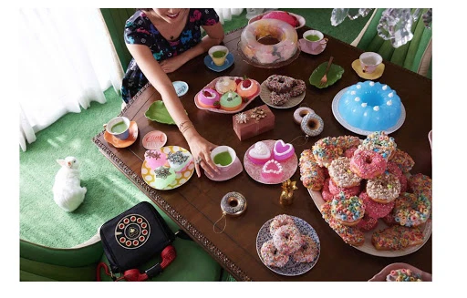 Overhead shot of woman at table with colorful donuts, cakes, and teacups, set indoors with pastel green carpet and a retro black telephone.