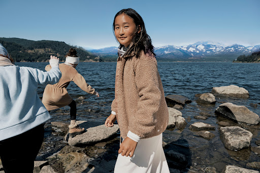 Smiling Asian girl in a sherpa jacket standing on rocks at a lake with two other children and snow-capped mountains in the background.