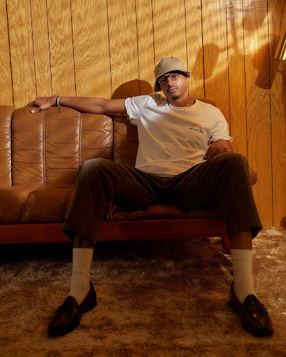 Adult male poses on a leather sofa in a casual t-shirt, trousers, and loafers, wearing glasses and a bucket hat against a wood-paneled wall.