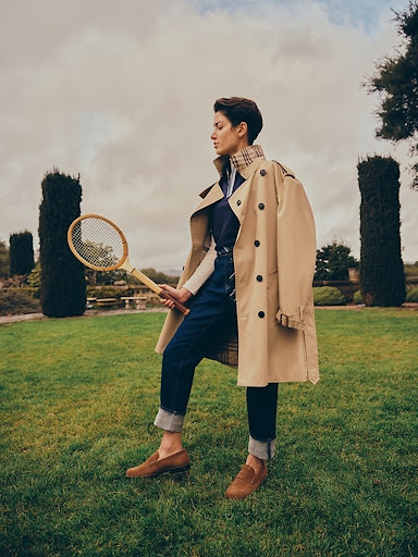 Woman in trench coat holds tennis racket on lawn with hedges and cloudy sky in background, showcasing fashion and outdoor lifestyle.