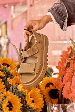 Tan UGG sandal held by a hand, styled with orange and yellow flowers. The footwear is the focus, showcasing its color and design.