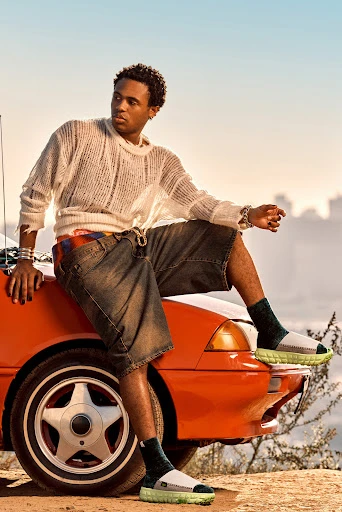 Young adult man poses on the hood of an orange car wearing casual clothes, sweater, and sandals in an outdoor setting with buildings.