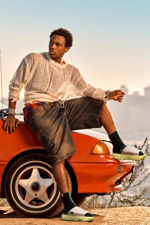 Young adult man poses on the hood of an orange car wearing casual clothes, sweater, and sandals in an outdoor setting with buildings.