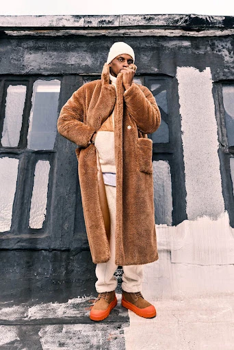 A Black male model showcases a long brown faux fur coat with tan pants and bright orange shoes against a textured, abstract backdrop.