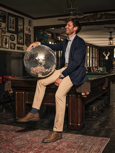 Smiling man in blazer and khaki pants sits on a pool table holding a disco ball, indoors with traditional decor and soft, warm lighting.