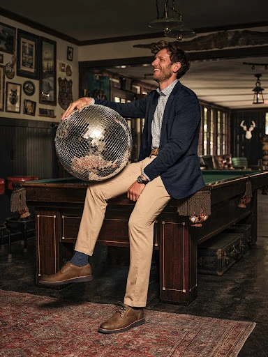 Smiling man in blazer and khaki pants sits on a pool table holding a disco ball, indoors with traditional decor and soft, warm lighting.